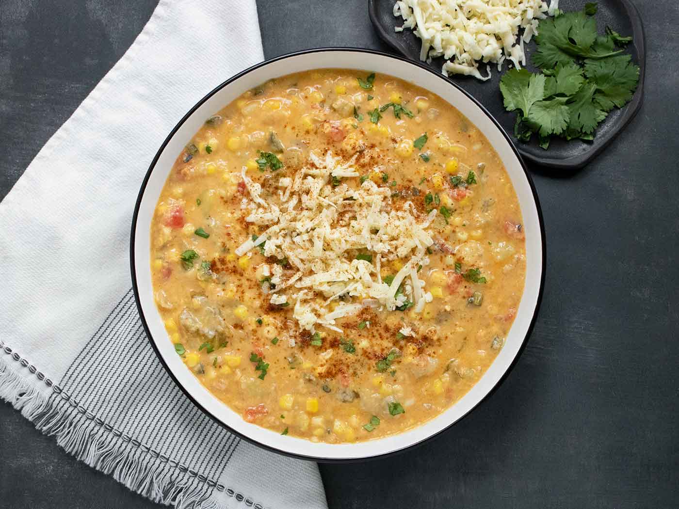 An overhead view of a bowl of Southwest Corn Chowder, with shredded cheese and cilantro on the side.