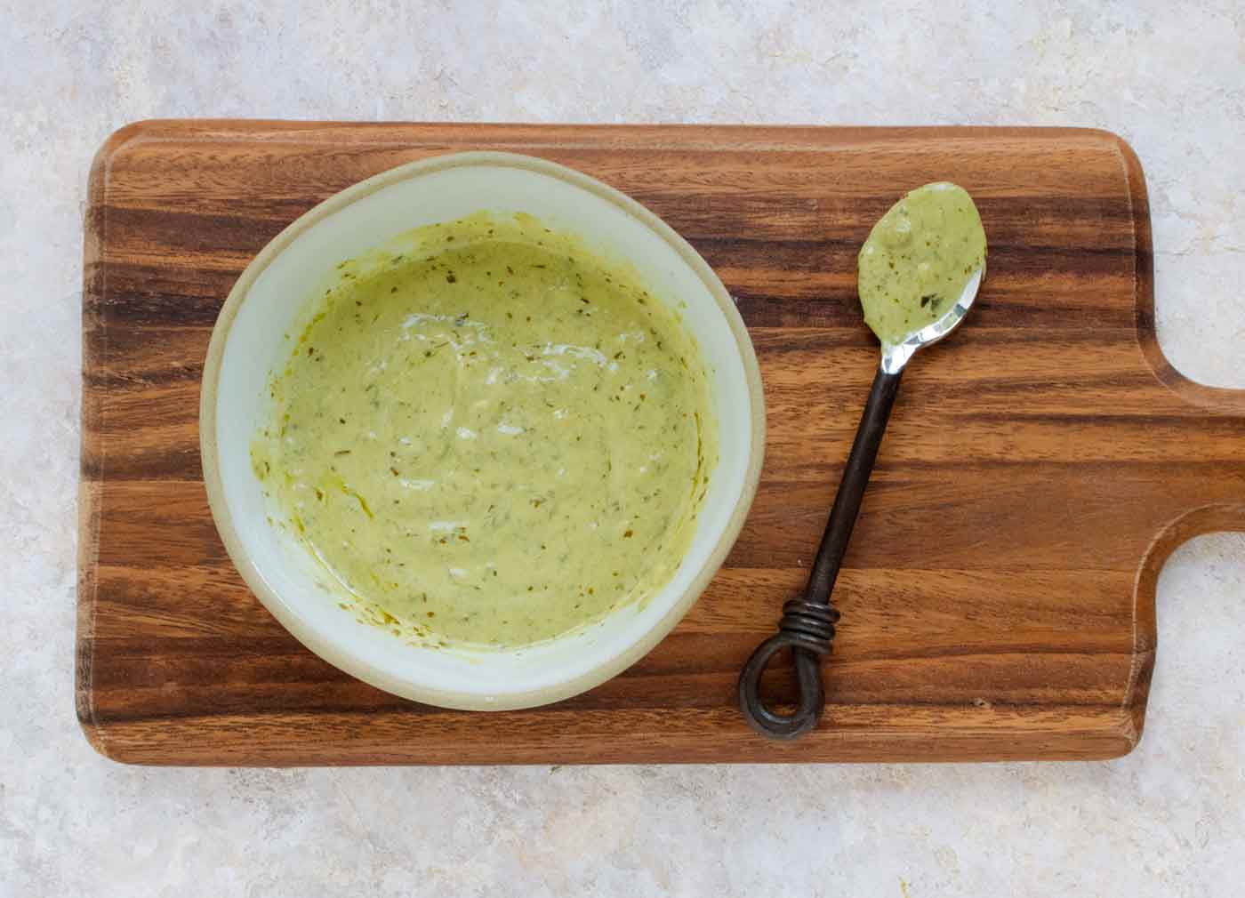 A bowl of creamy pesto dressing on a wooden serving board, with a small serving spoon.